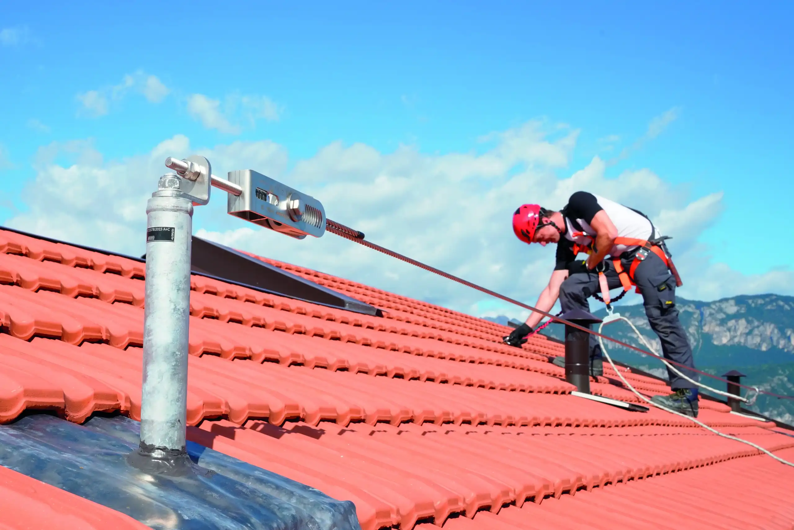 Foto di un installatore di un pannello fotovoltaico sopra un tetto agganciato ad un palo con una corda di sicurezza di Rothoblass. Nella parte anteriore della foto si vede la parte superiore del tetto rosso di un'abitazioe con sullo sfondo il cielo azzurro